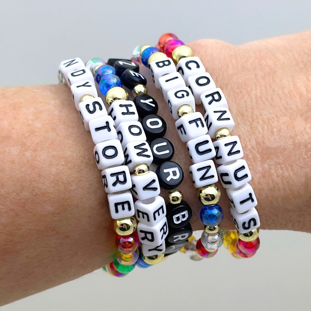 Broadway Stacks CORN NUTS Collection. 5 stretch bracelets included in Stack. Red, yellow, green, blue, black and gold colored beads. Letter beads that spell quotes. Gold star logo bead to remind you that you shine. Heathers the Musical fans.