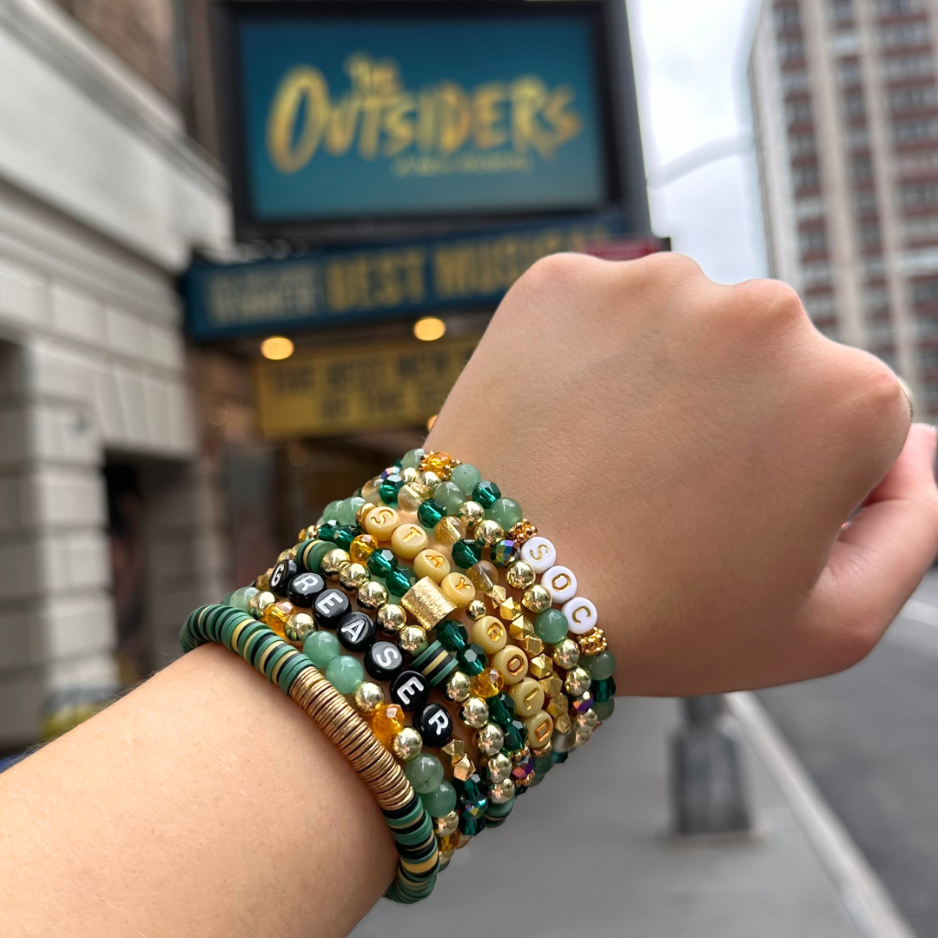 Broadway Stacks STAY GOLD collection. Stretch bracelets included in Stack. Gold, yellow, green and grey colored beads. Letter beads that have show quote or name. Broadway Stacks gold star logo bead to remind you that You Shine. The Outsiders the Musical fans.