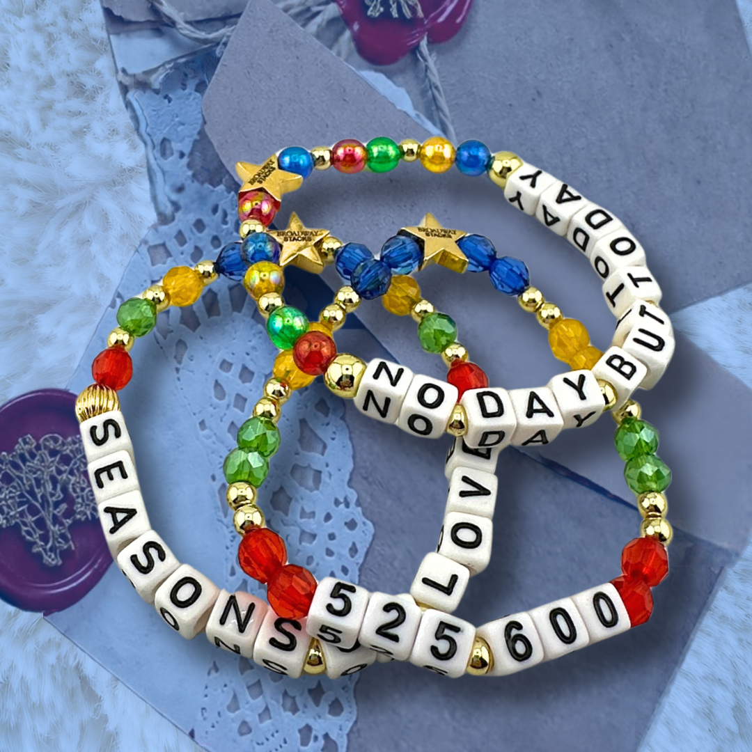 Broadway Stacks theatre-themed bracelets inspired by RENT the Musical. Three bracelets with quotes SEASONS OF LOVE, 525,600 and NO DAY BUT TODAY with red, green, yellow and blue beads on a love note with blue overlay.