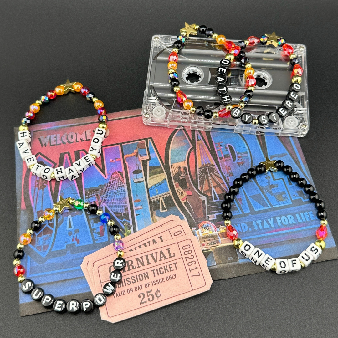 Broadway Stacks VAMPIRE Collection bead bracelets on a Santa Carla postcard. Black, red, orange and gold colored beads with letter beads spelling out quotes. Inspired by The Lost Boys Musical. Carnival tickets and cassette tape displayed also.
