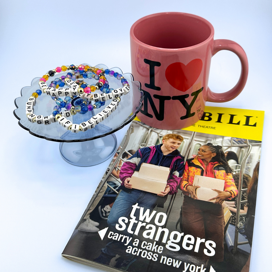 Pink mug with 'I ❤️ NY' text, cake stand with letter beads, and Playbill titled 'Two Strangers Carry a Cake Across New York' on a light blue background. Broadway Stacks CAKE Collection.