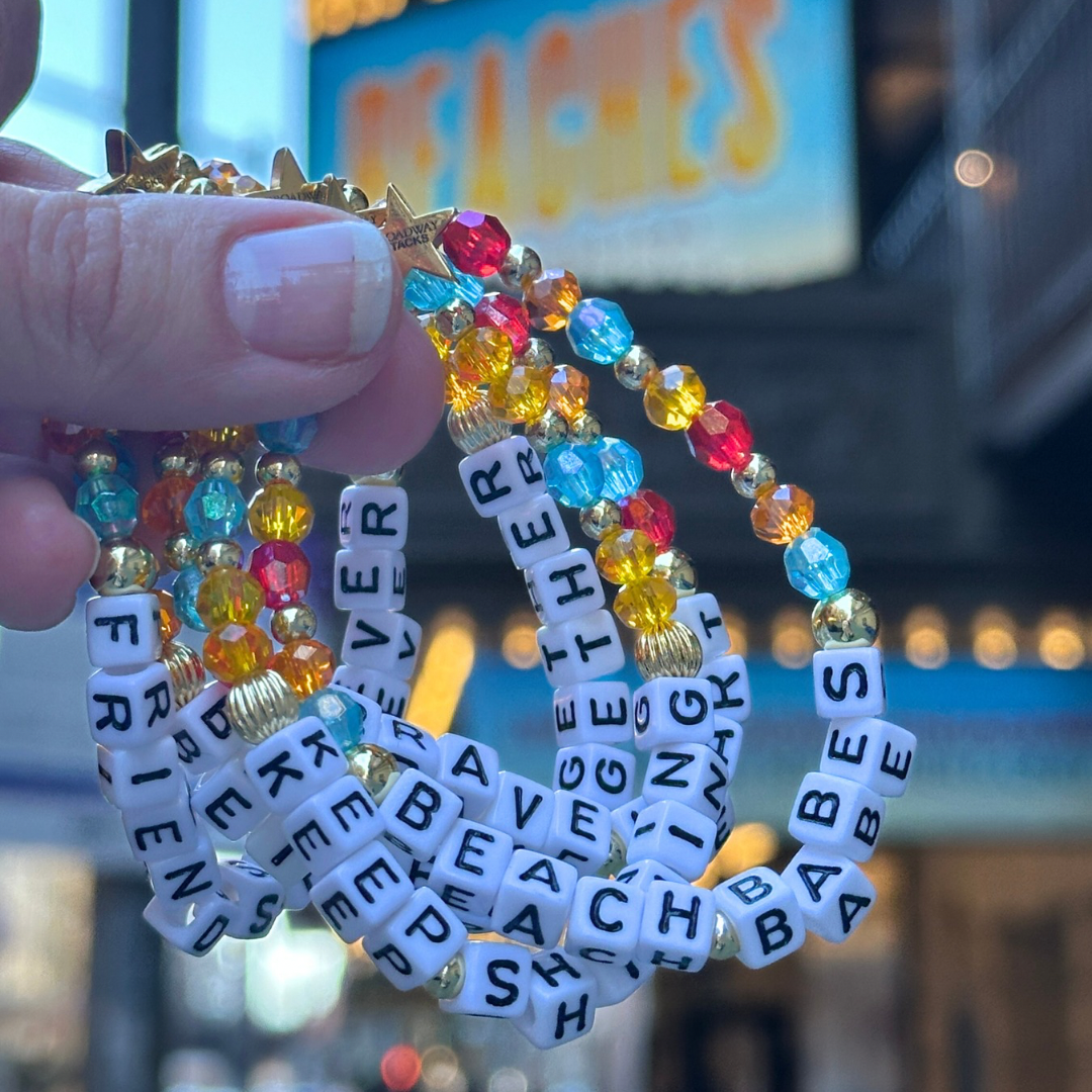 Broadway Stacks theatre-themed bracelets BETTER TOGETHER Collection inspired by Beaches the Musical. Blue yellow orange and red beads with gold-colored accents spell FRIENDS FOREVER, BEACH BABES. Blurred Broadway Beaches theater marquee in background.