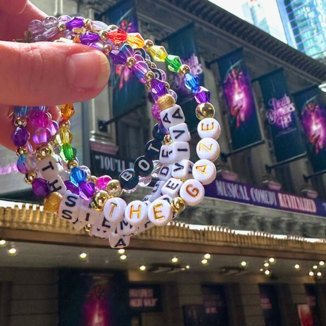 Broadway Stacks theatre-themed bracelet SIEMPRE VIVA Collection inspired by Death Becomes Her Musical. Purple, rainbow and gold beads. Letter beads that spell SIEMPRE VIVA, THE GAZE, THAT WAS RUDE & BOTTOMS UP. Bracelets in front of Broadway marquee.  