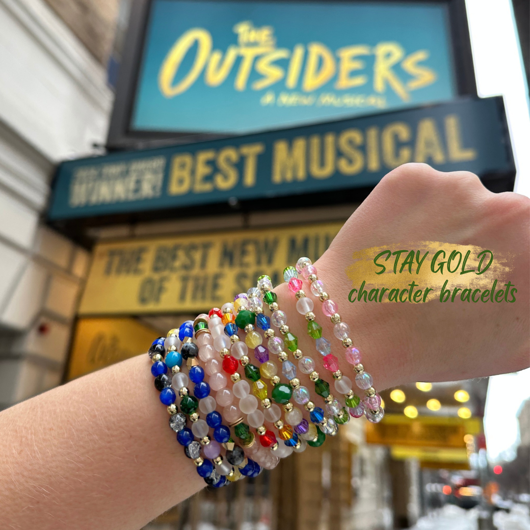 Broadway Stacks STAY GOLD character bracelets on wrist on front of The Outsiders Musical marquee