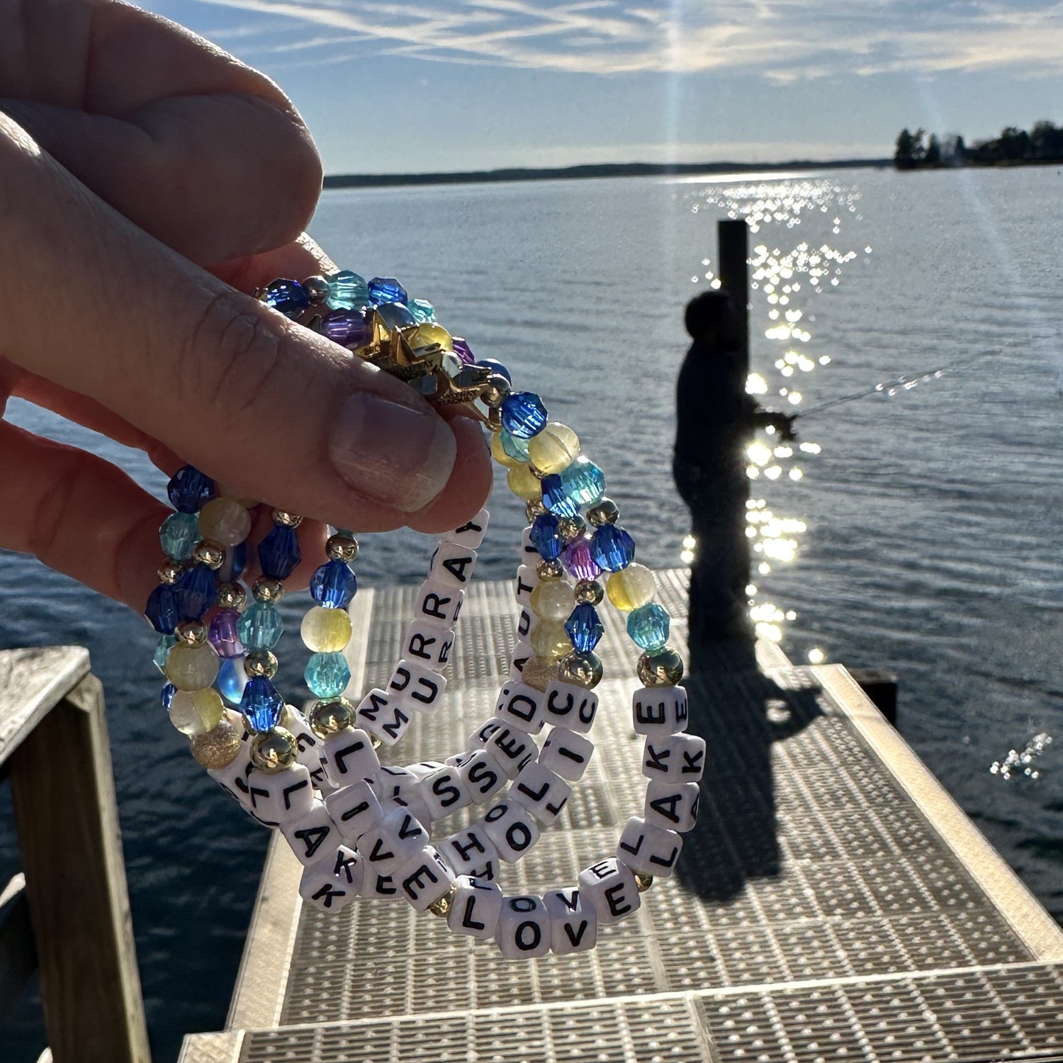 Broadway Stacks bead bracelets LAKE Collection. Man on dock fishing with sun on water. Hand holding bracelets in yellow and blue colors.