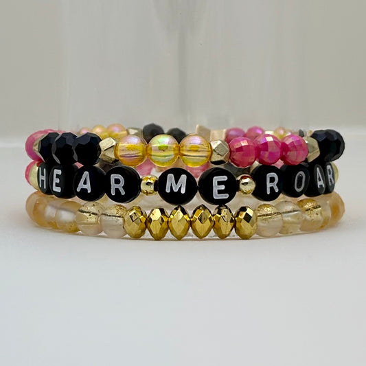 Broadway Stacks De Bois Band collection. 3 stretch bracelets included in Stack. Pinks, golds and black colored beads. Letter beads that spell Hear Me Roar. Broadway Stacks gold star logo bead on back.