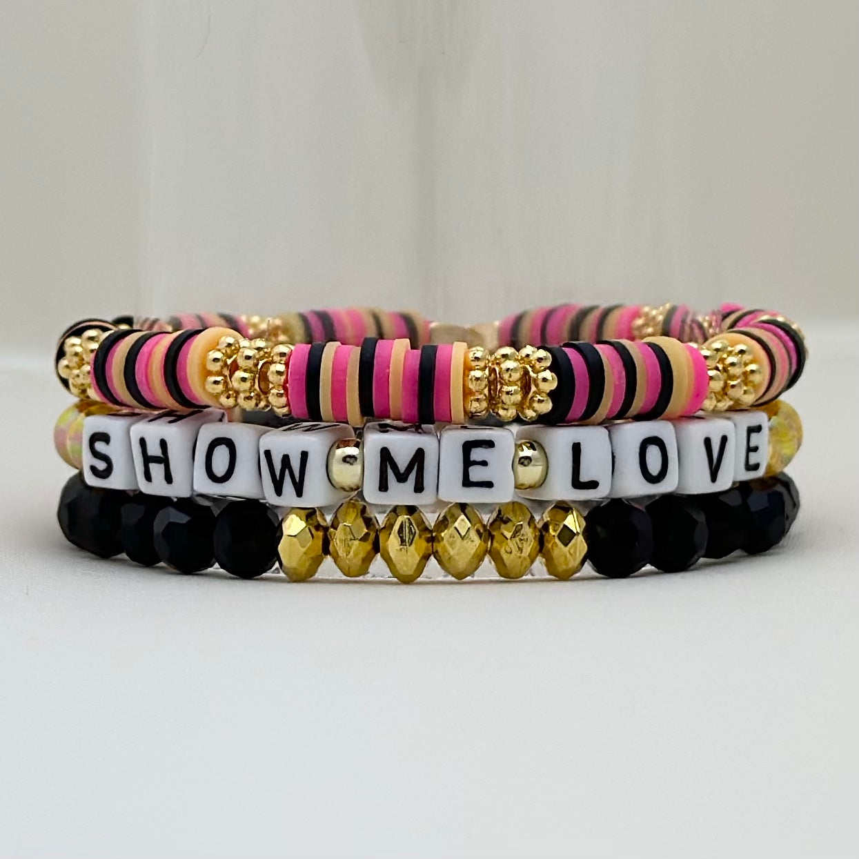 Broadway Stacks De Bois Band collection. 3 stretch bracelets included in Stack. Pinks, golds and black colored beads. Letter beads that spell Show Me Love. Broadway Stacks gold star logo bead on back.