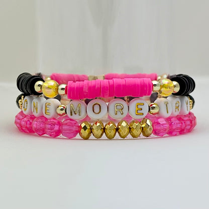 Broadway Stacks De Bois Band collection. 3 stretch bracelets included in Stack. Pinks, golds and black colored beads. Letter beads that spell One More Try. Broadway Stacks gold star logo bead on back.