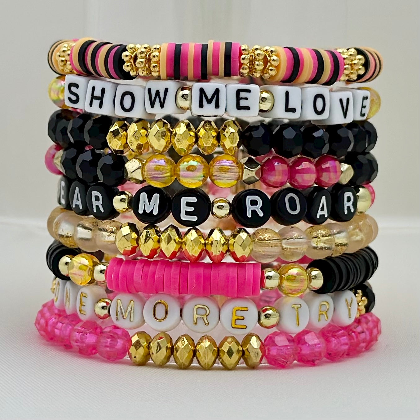 Broadway Stacks De Bois Band collection. 3 stretch bracelets included in Stack. Pinks, golds and black colored beads. Letter beads that spell One More Try. Broadway Stacks gold star logo bead on back.