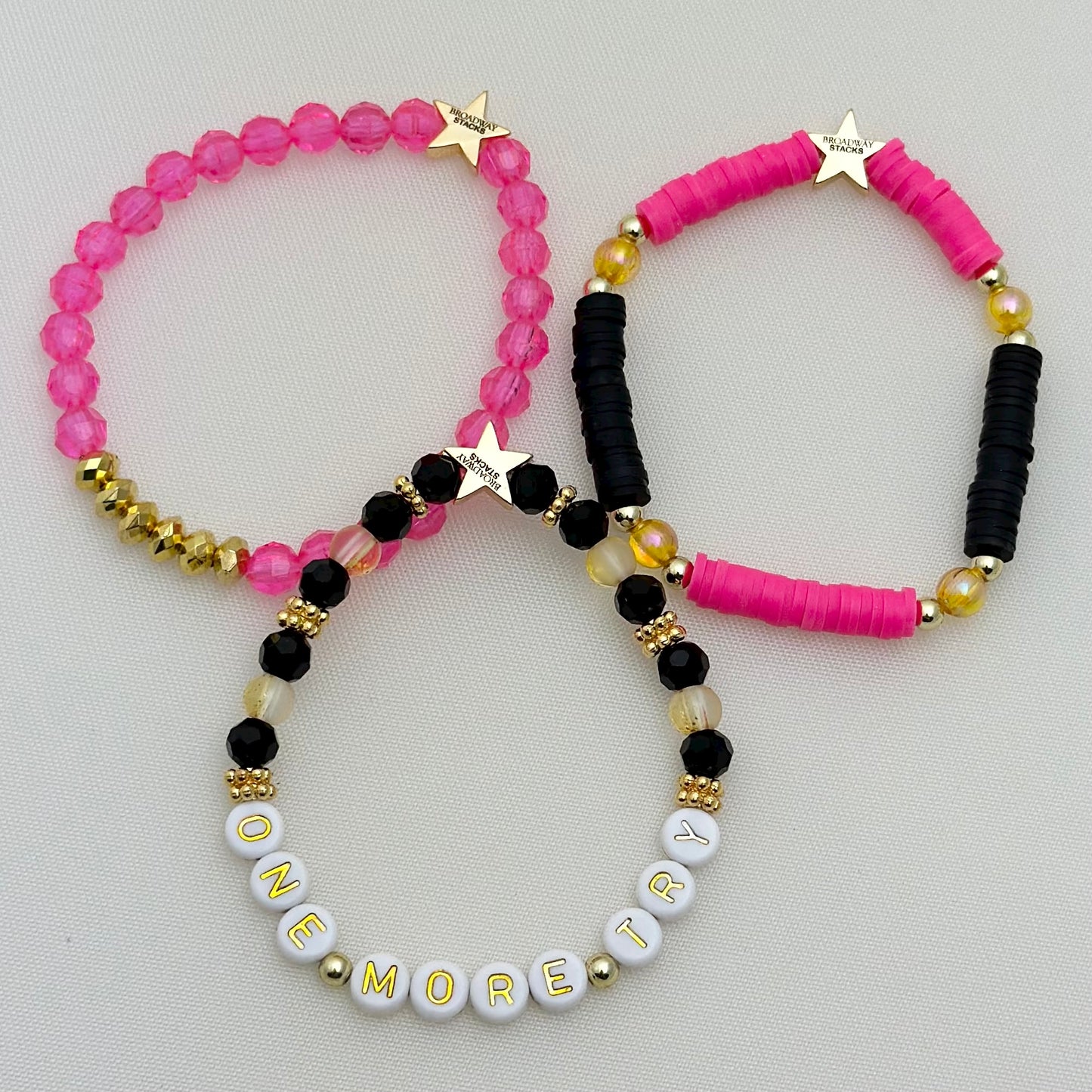 Broadway Stacks De Bois Band collection. 3 stretch bracelets included in Stack. Pinks, golds and black colored beads. Letter beads that spell One More Try. Broadway Stacks gold star logo bead on back.