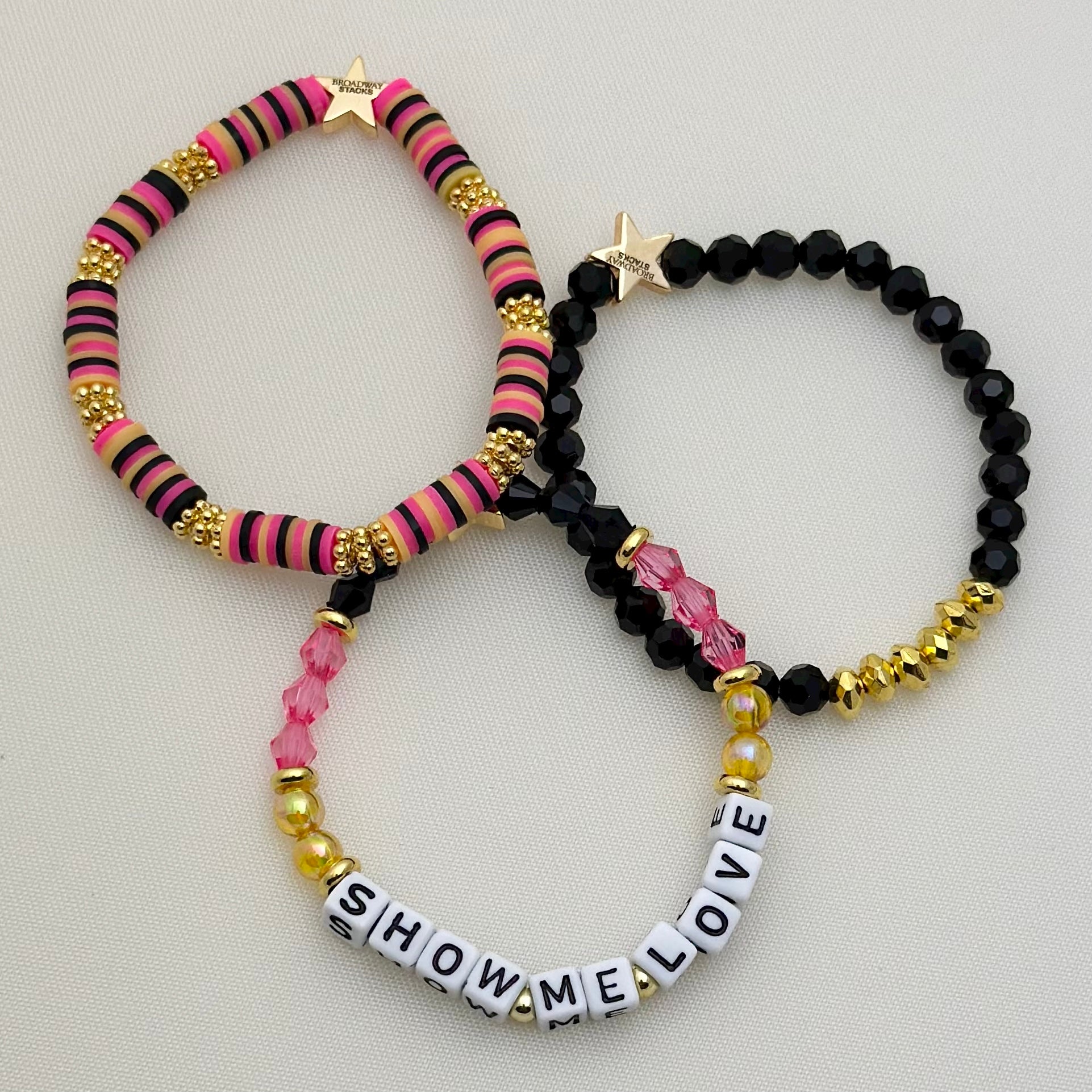 Broadway Stacks De Bois Band collection. 3 stretch bracelets included in Stack. Pinks, golds and black colored beads. Letter beads that spell Show Me Love. Broadway Stacks gold star logo bead on back.