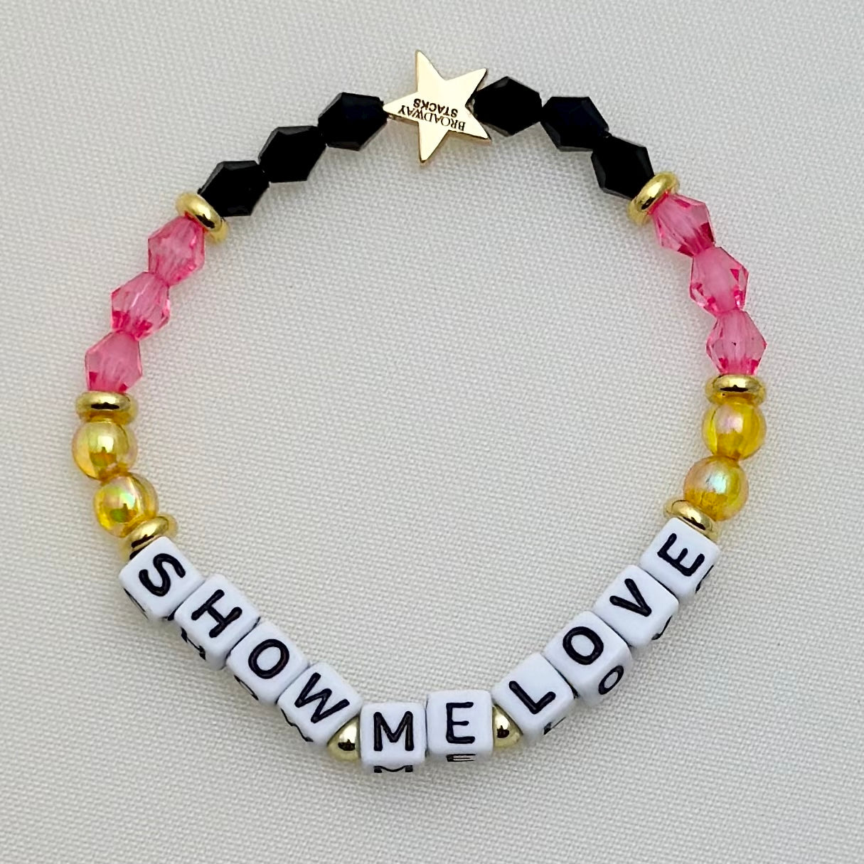 Broadway Stacks De Bois Band collection. 3 stretch bracelets included in Stack. Pinks, golds and black colored beads. Letter beads that spell Show Me Love. Broadway Stacks gold star logo bead on back.