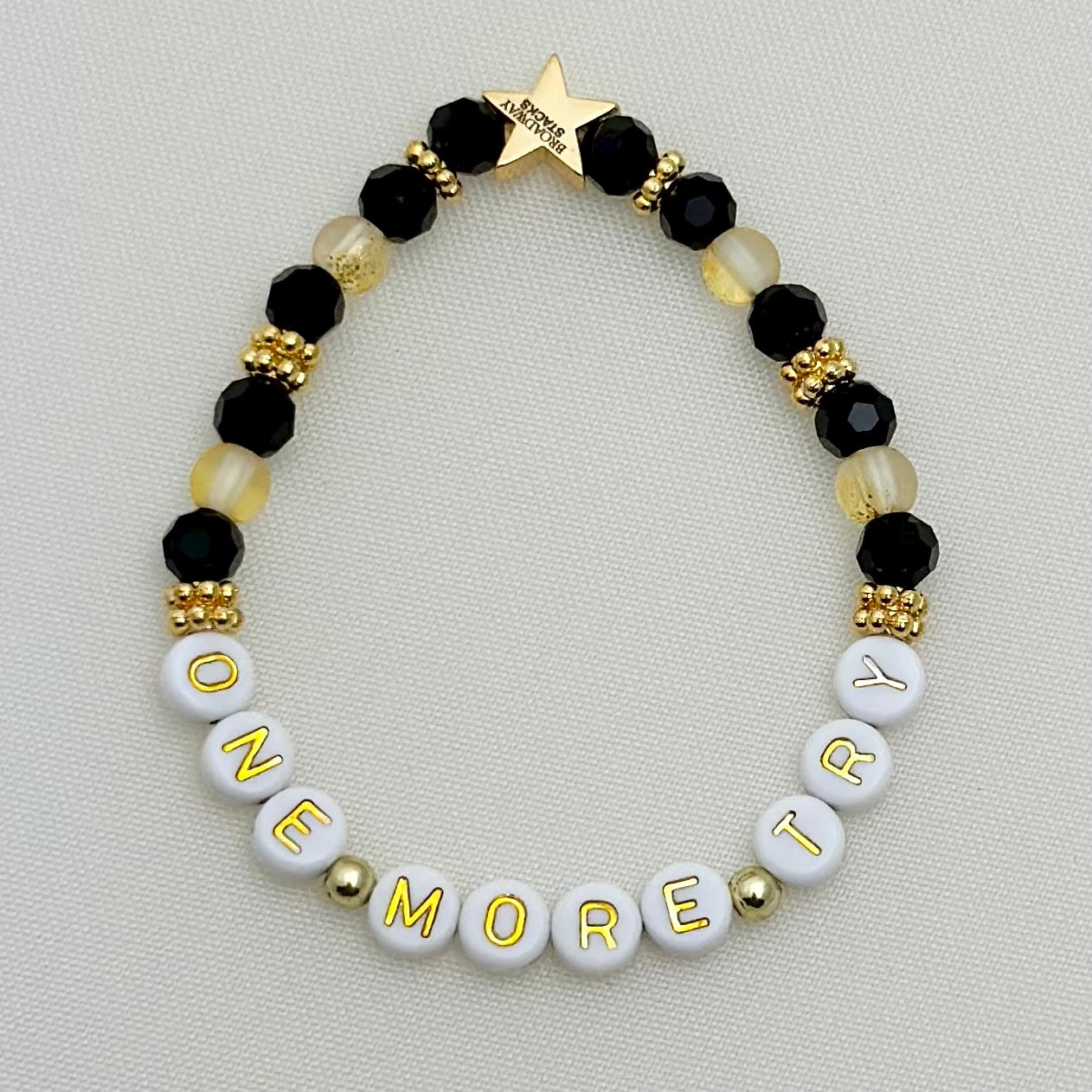 Broadway Stacks De Bois Band collection. 3 stretch bracelets included in Stack. Pinks, golds and black colored beads. Letter beads that spell One More Try. Broadway Stacks gold star logo bead on back.