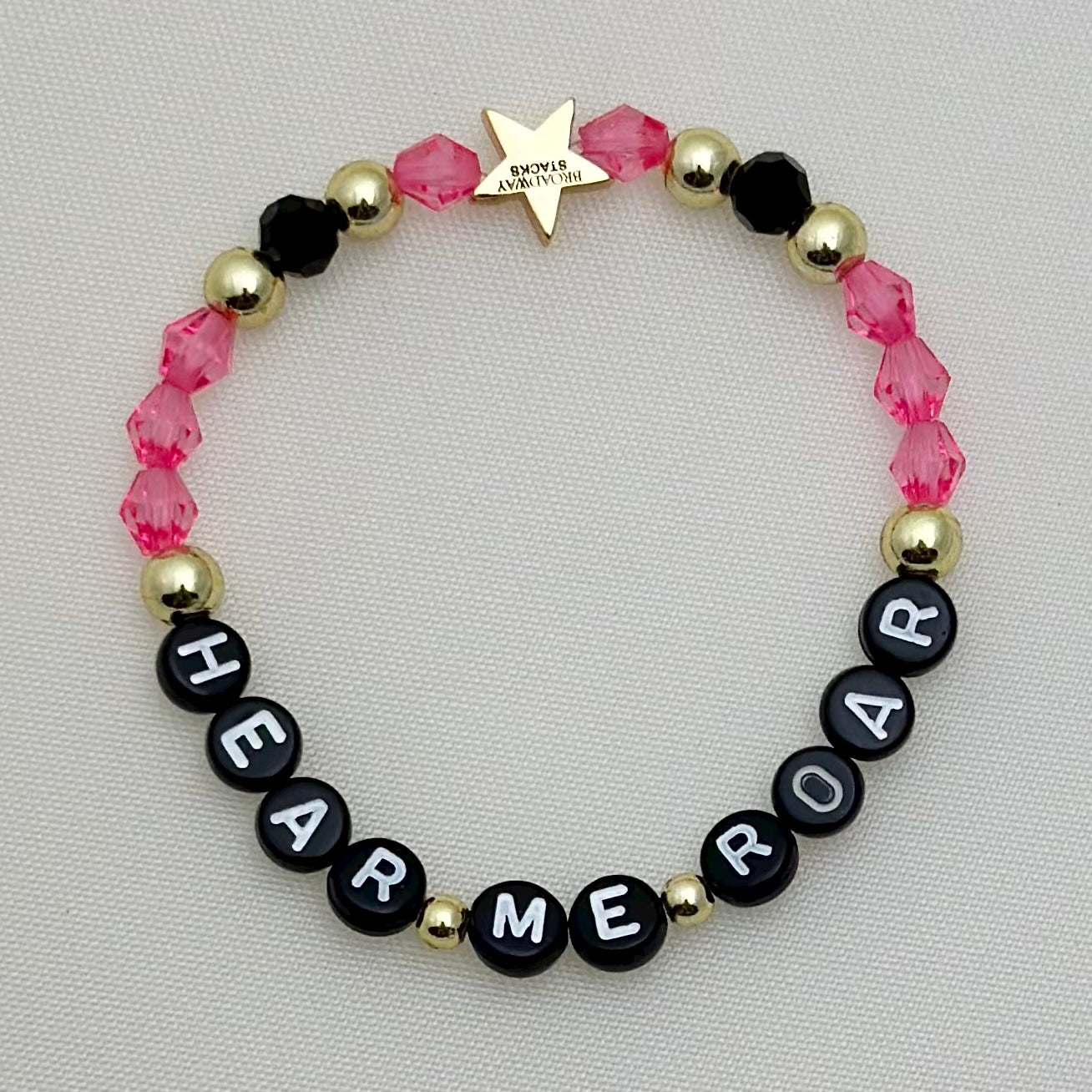 Broadway Stacks De Bois Band collection. 3 stretch bracelets included in Stack. Pinks, golds and black colored beads. Letter beads that spell Hear Me Roar. Broadway Stacks gold star logo bead on back.