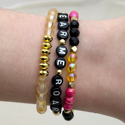 Broadway Stacks De Bois Band collection. 3 stretch bracelets included in Stack. Pinks, golds and black colored beads. Letter beads that spell Hear Me Roar. Broadway Stacks gold star logo bead on back.