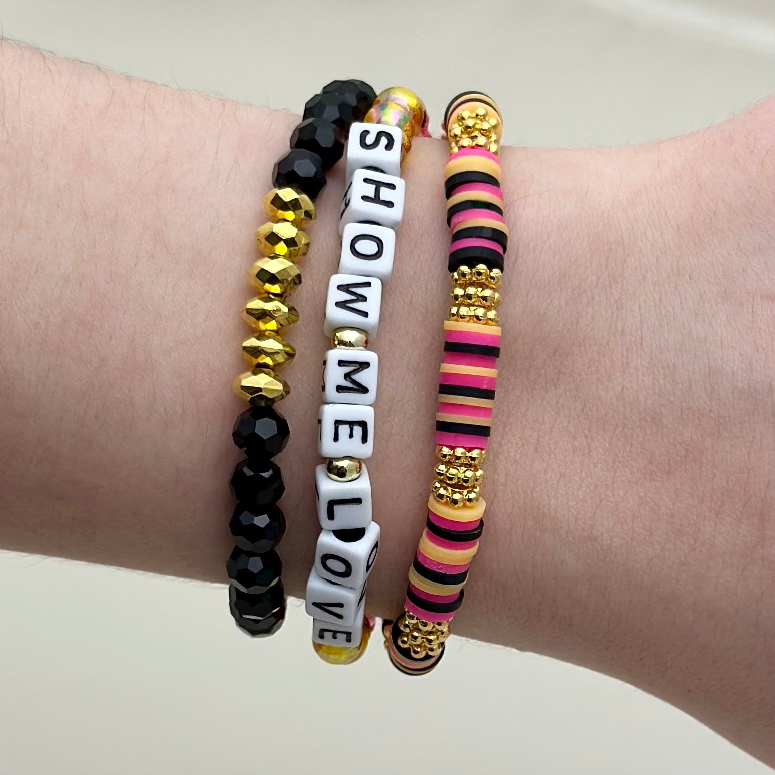 Broadway Stacks De Bois Band collection. 3 stretch bracelets included in Stack. Pinks, golds and black colored beads. Letter beads that spell Show Me Love. Broadway Stacks gold star logo bead on back.