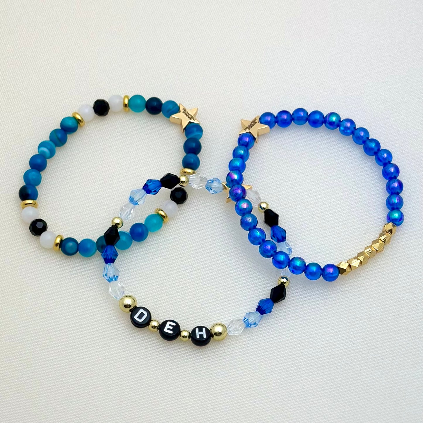 Broadway Stacks SKY collection. 3 stretch bracelets included in Stack. Blues, white, black and gold colored beads. Letter beads that spell DEH.