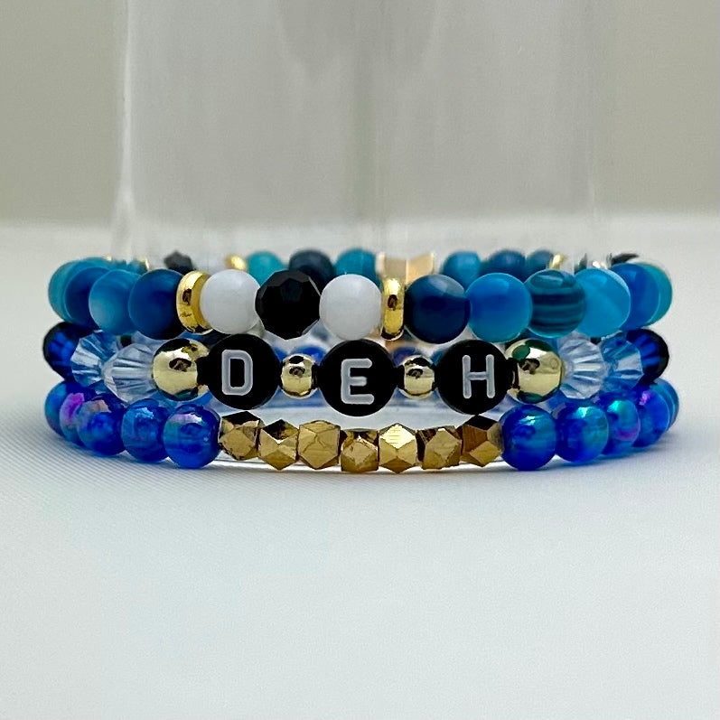 Broadway Stacks SKY collection. 3 stretch bracelets included in Stack. Blues, white, black and gold colored beads. Letter beads that spell DEH.