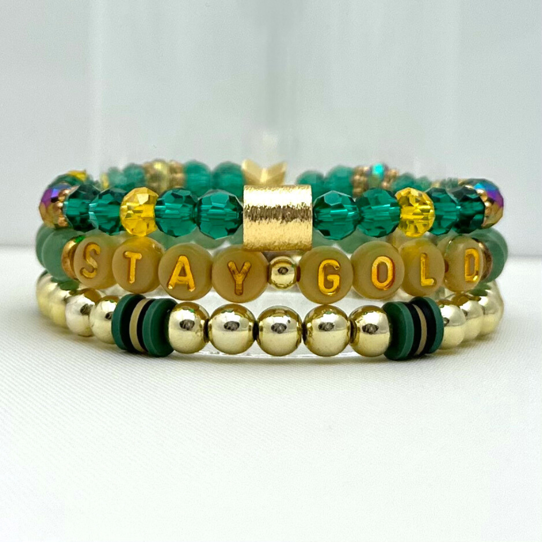 Broadway Stacks STAY GOLD collection. Stretch bracelets included in Stack. Gold, yellow, green and grey colored beads. Letter beads that have show quote or name. Broadway Stacks gold star logo bead on back.