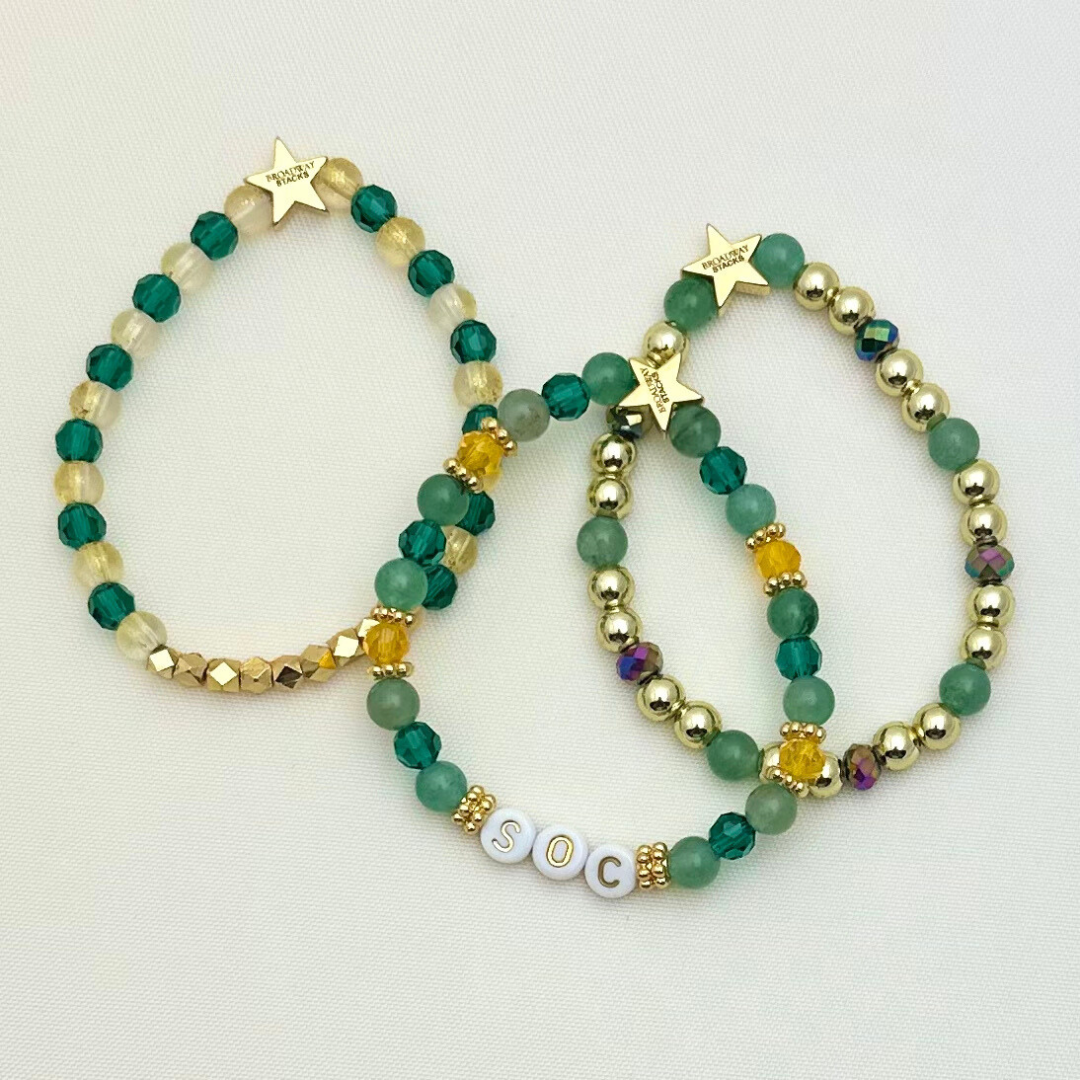 Broadway Stacks STAY GOLD collection. Stretch bracelets included in Stack. Gold, yellow, green and grey colored beads. Letter beads that have show quote or name. Broadway Stacks gold star logo bead on back.