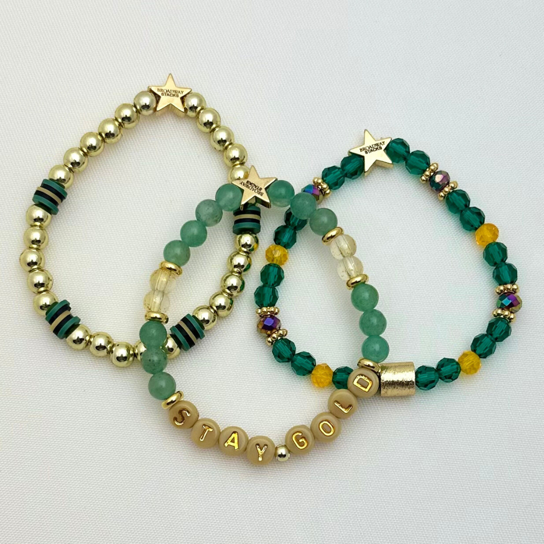 Broadway Stacks STAY GOLD collection. Stretch bracelets included in Stack. Gold, yellow, green and grey colored beads. Letter beads that have show quote or name. Broadway Stacks gold star logo bead on back.