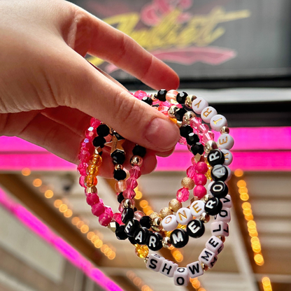 Broadway Stacks De Bois Band collection. Pink, yellow, gold and black colored beads on bracelets. Background is & Juliet the Musical marquee in NYC.