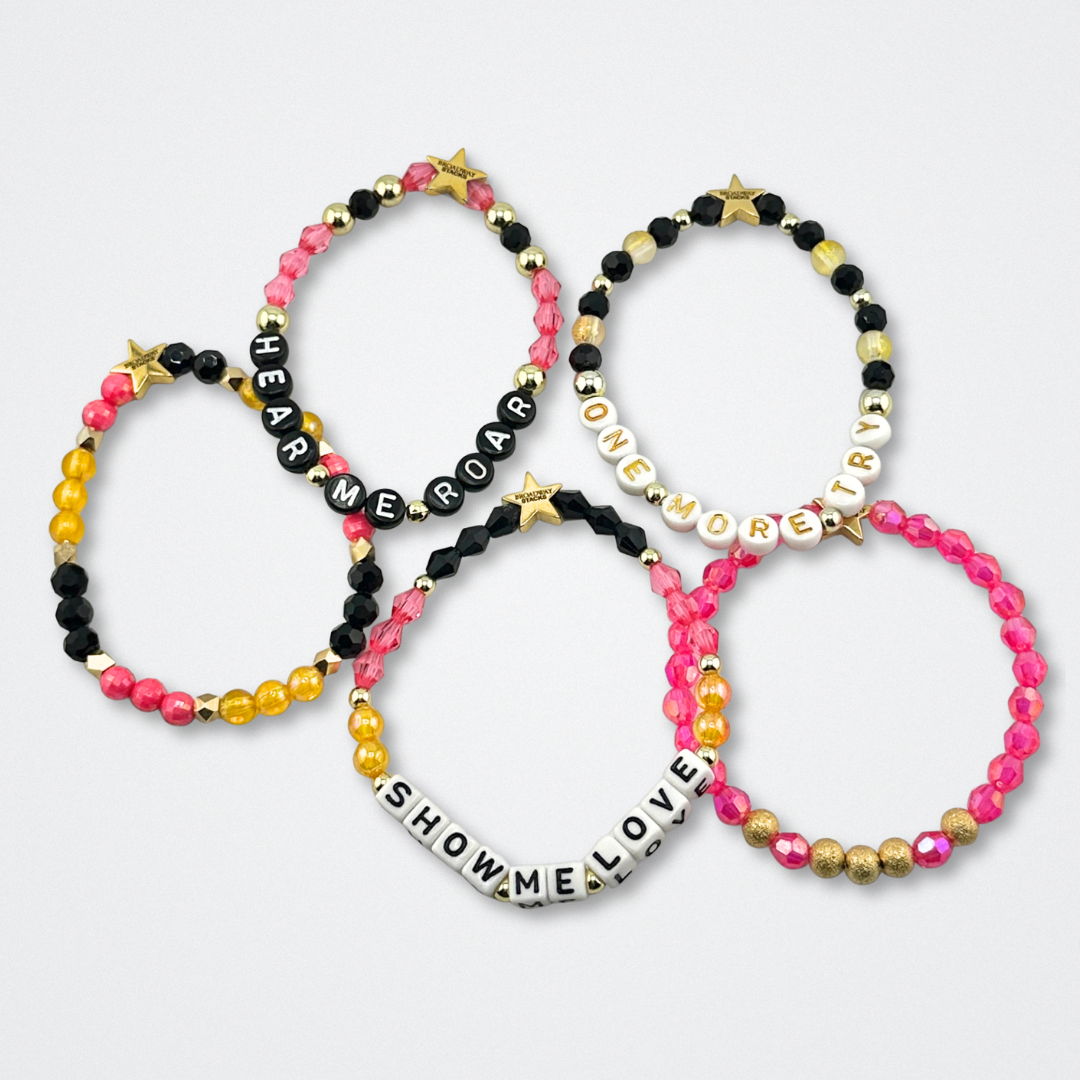 Broadway Stacks De Bois Band collection. Pink, yellow, gold and black colored beads on bracelets. Background is gray. Inspired by & Juliet the Musical.