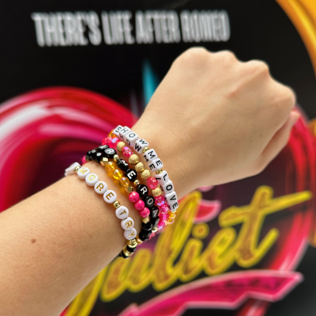 Broadway Stacks De Bois Band collection. Pink, yellow, gold and black colored beads on bracelets. Arm wearing bracelets on wrist with the background of & Juliet the Musical marquee in NYC.