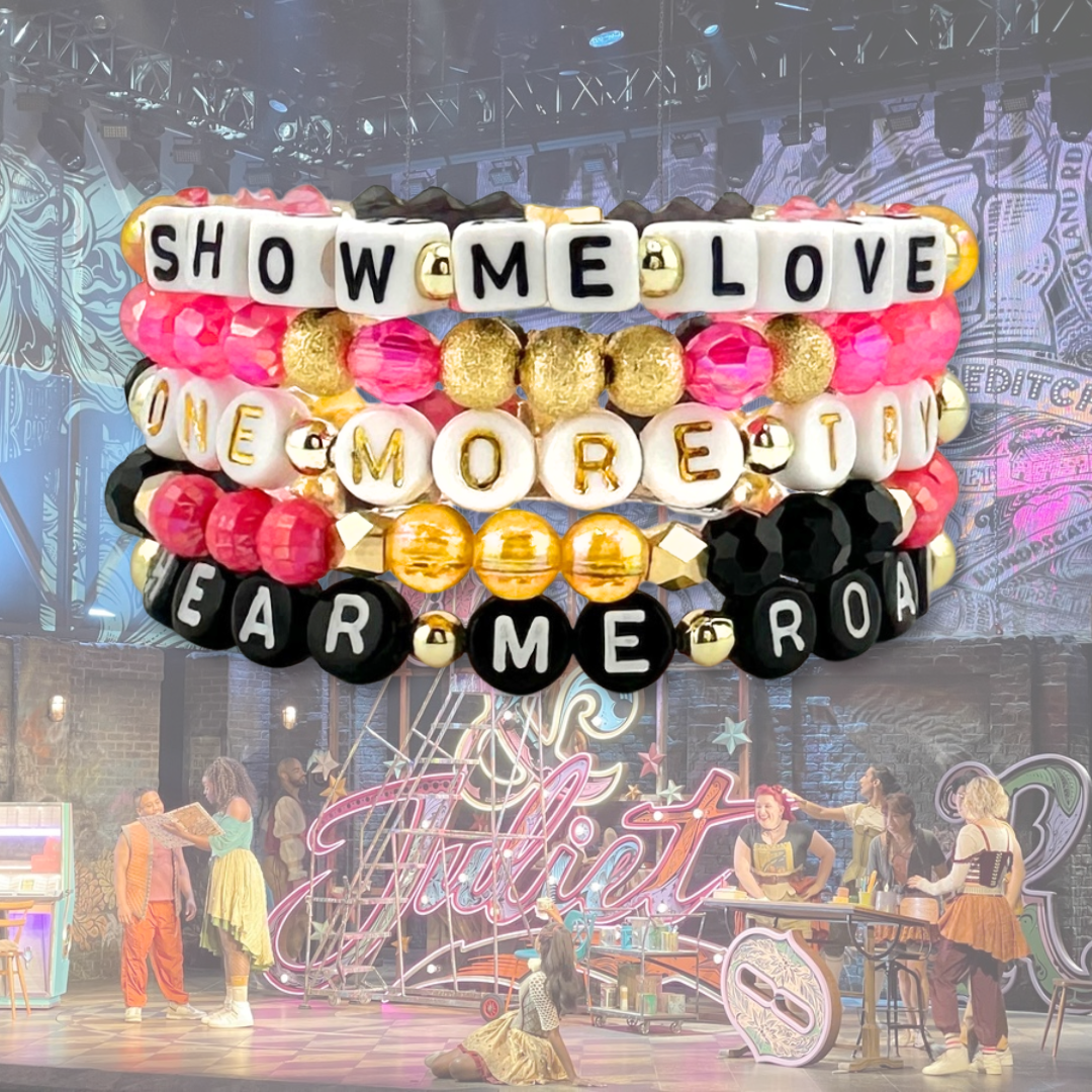 Broadway Stacks De Bois Band collection. Bracelets stacked with pink, yellow, gold and black colored beads and letter beads. Background is & Juliet the Musical set on Broadway in NYC.