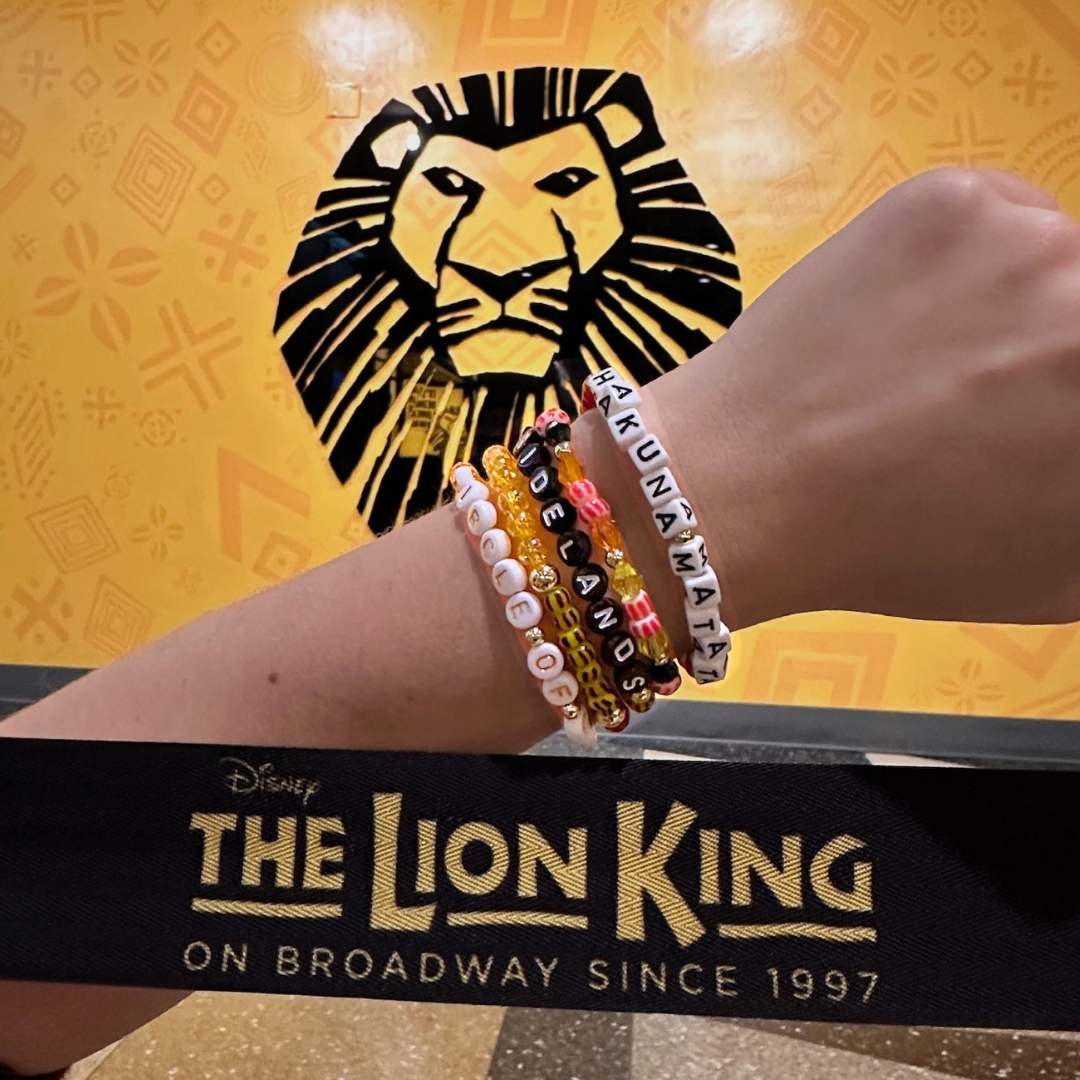 Broadway Stacks SAFARI collection.Yellow, orange, red, black, animal print and gold colored beads on bracelets. Wrist wearing bracelets stacked in front of The Lion King on Broadway in NYC.