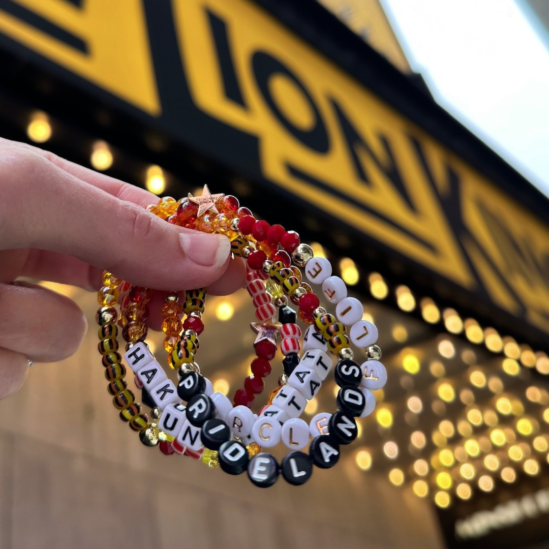 Broadway Stacks SAFARI collection.Yellow, orange, red, black, animal print and gold colored beads on bracelets. Hand holding bracelets stacked in front of The Lion King on Broadway marquee in NYC.