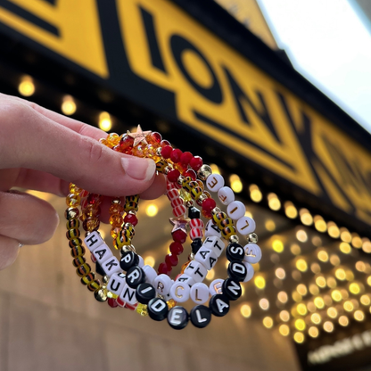 Broadway Stacks SAFARI collection.Yellow, orange, red, black, animal print and gold colored beads on bracelets. Hand holding bracelets stacked in front of The Lion King on Broadway marquee in NYC.