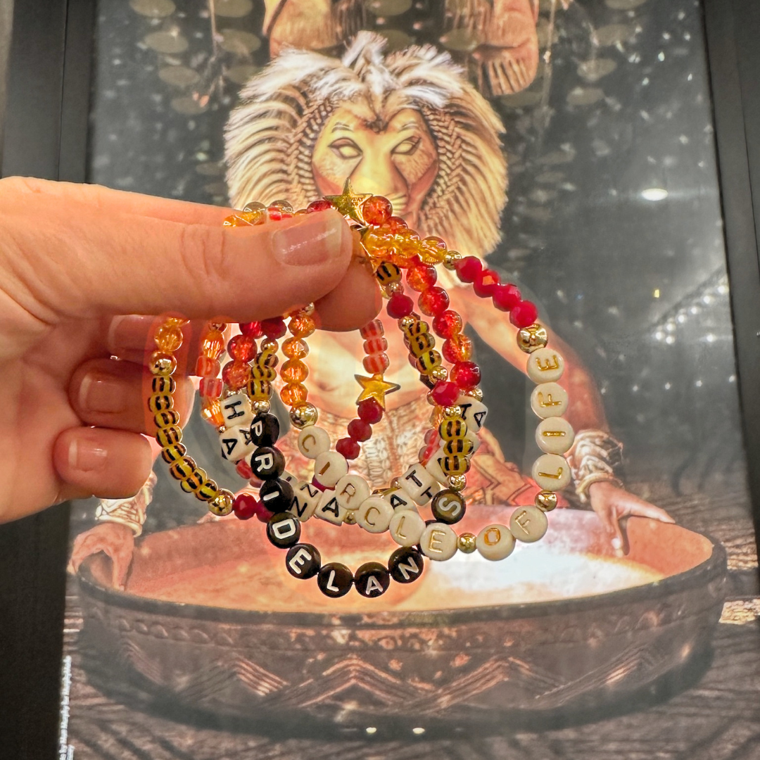 Broadway Stacks SAFARI collection.Yellow, orange, red, black, animal print and gold colored beads on bracelets. Hand holding bracelets stacked in front of The Lion King on Broadway marquee at the Marriott in NYC.