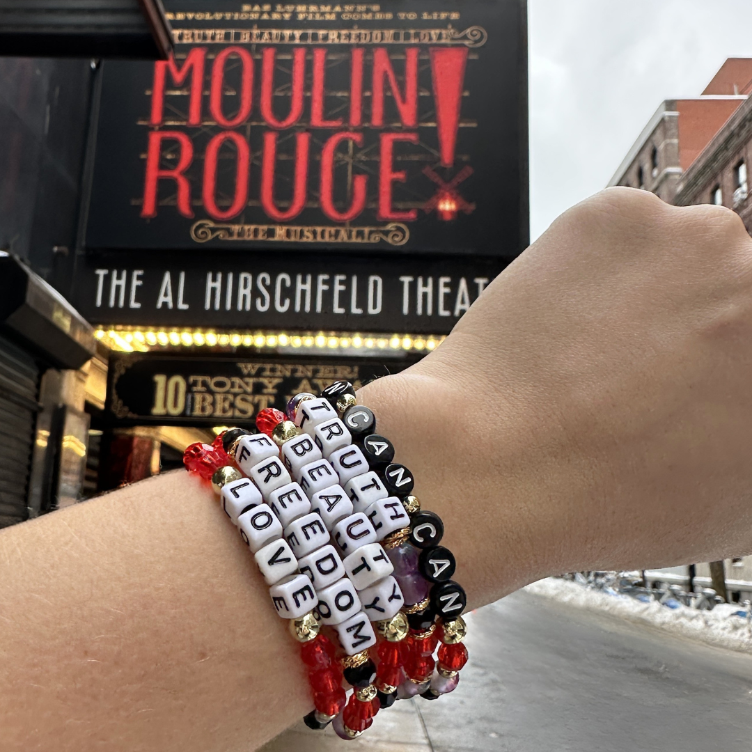 Broadway Stacks RED LIGHT bracelets on wrist in front of theatre marquee for Moulin Rouge, Al Hirschfeld Theatre