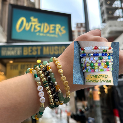 Broadway Stacks STAY GOLD bracelets on a wrist. the STAY GOLD Character-inspired bracelets in a smaller photo. Both in front of The Outsiders Musical marquee/.