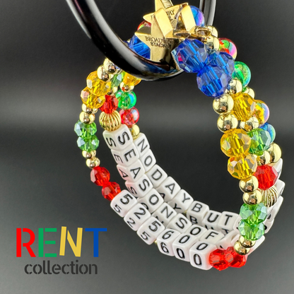 Broadway Stacks theatre-themed bracelets inspired by RENT the Musical. Three bracelets with quotes SEASONS OF LOVE, 525,600 and NO DAY BUT TODAY with red, green, yellow and blue beads hanging with black background. Signature star bead to remind you that YOU SHINE!