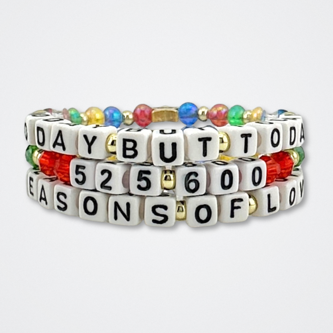 Broadway Stacks theatre-themed bracelets inspired by RENT the Musical. Three-stack of bracelets with quotes SEASONS OF LOVE, 525,600 and NO DAY BUT TODAY with red, green, yellow and blue beads on a gray background. 