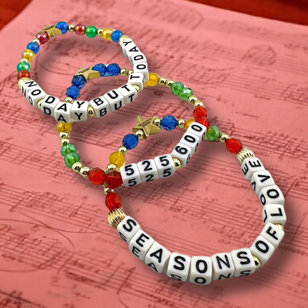 Broadway Stacks theatre-themed bracelets inspired by RENT the Musical. Three bracelets with quotes SEASONS OF LOVE, 525,600 and NO DAY BUT TODAY with red, green, yellow and blue beads on a piece of sheet music with red overlay.