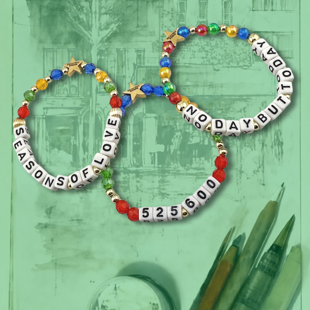 Broadway Stacks theatre-themed bracelets inspired by RENT the Musical. Three bracelets with quotes SEASONS OF LOVE, 525,600 and NO DAY BUT TODAY with red, green, yellow and blue beads on an artist's canvas with green overlay.