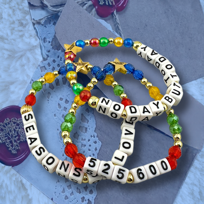 Broadway Stacks theatre-themed bracelets inspired by RENT the Musical. Three bracelets with quotes SEASONS OF LOVE, 525,600 and NO DAY BUT TODAY with red, green, yellow and blue beads on a love note with blue overlay.