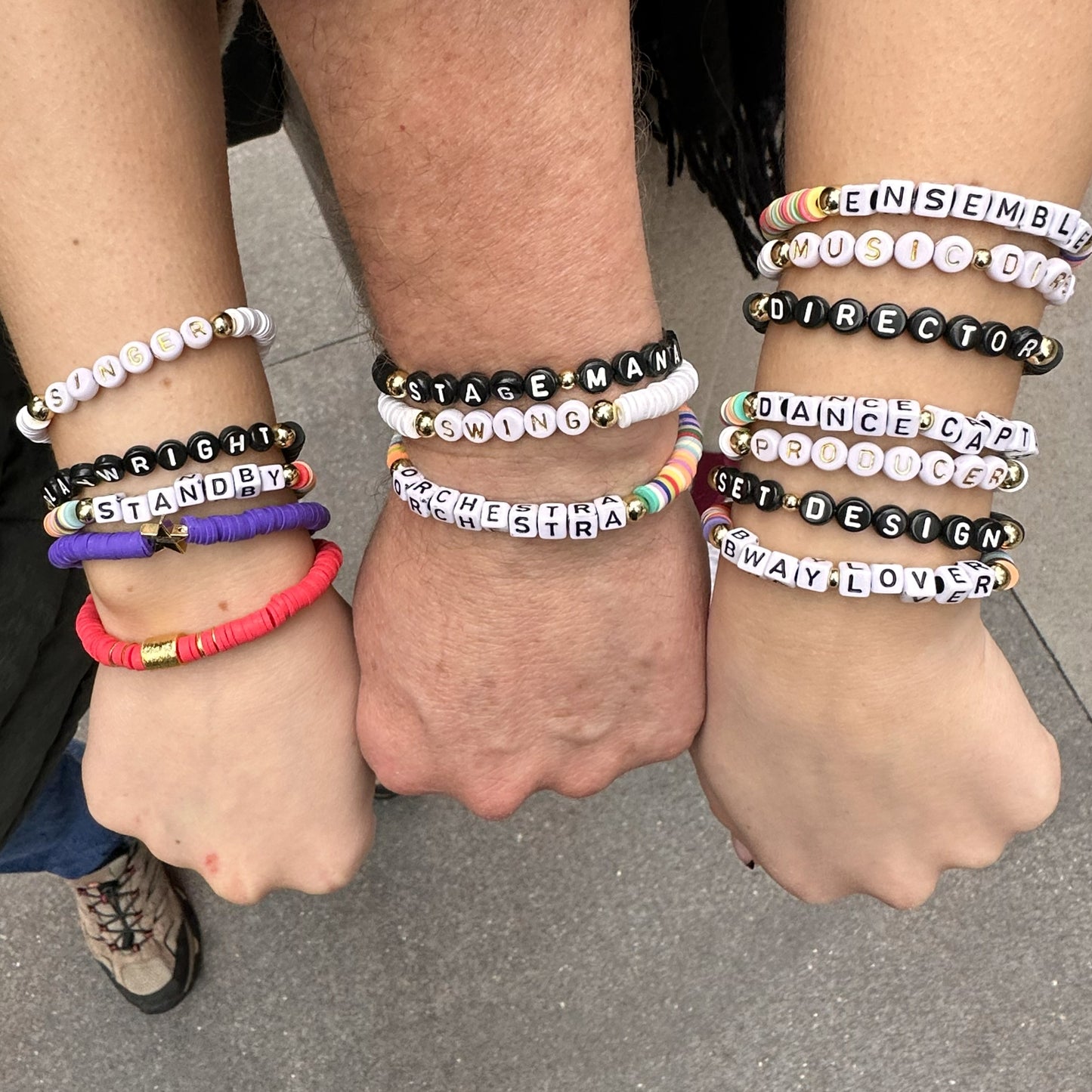 Three wrists wearing multiple bracelets with various text. Broadway Stacks theatre-themed Spotlight bracelets highlighting ALL theater roles. Because everyone deserves to shine.