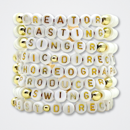 Bracelet with white beads featuring gold letters on a white background. Broadway Stacks theatre-themed Spotlight bracelets highlighting ALL theater roles. Because everyone deserves to shine.