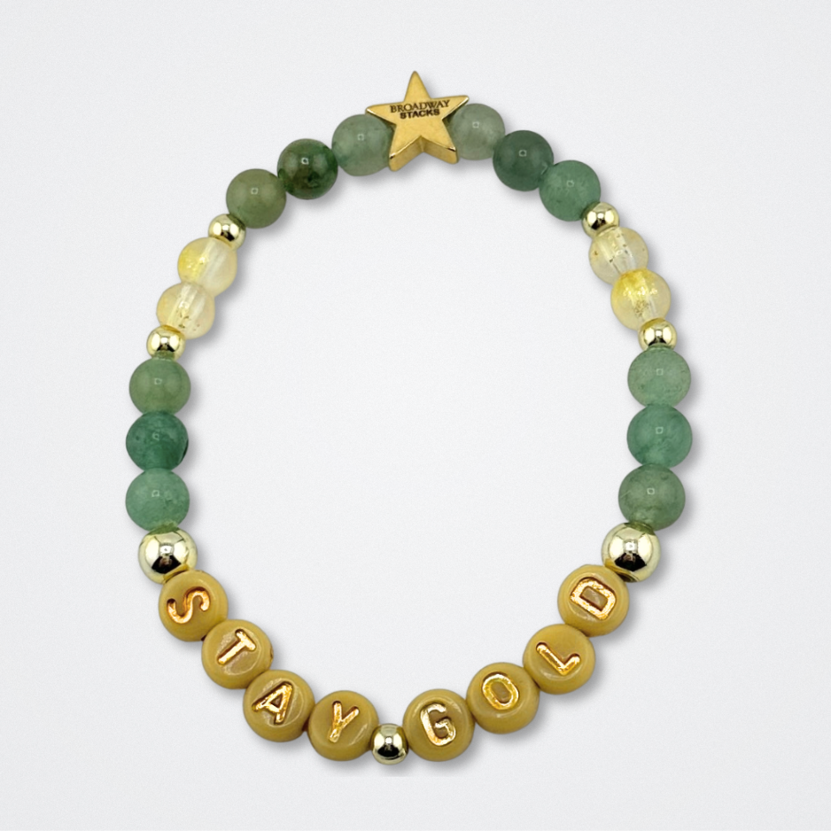 Broadway Stacks STAY GOLD bracelet inspired by The Outsiders Musical. Green and gold beads with letter beads that spell STAY GOLD. Logo star bead to remind you that YOU SHINE. Displayed on a gray background.