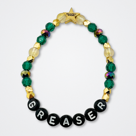 Broadway Stacks STAY GOLD bracelet inspired by The Outsiders Musical. Green and gold beads with letter beads that spell GREASER. Logo star bead to remind you that YOU SHINE. Displayed on a gray background.
