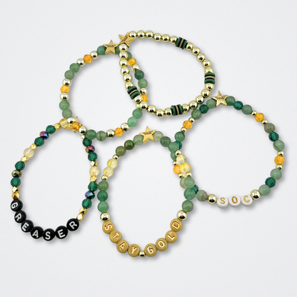 Broadway Stacks STAY GOLD bracelets inspired by The Outsiders Musical. Green, yellow and gold beads with letter beads that spell quotes. Logo star bead to remind you that YOU SHINE. Displayed on a gray background.
