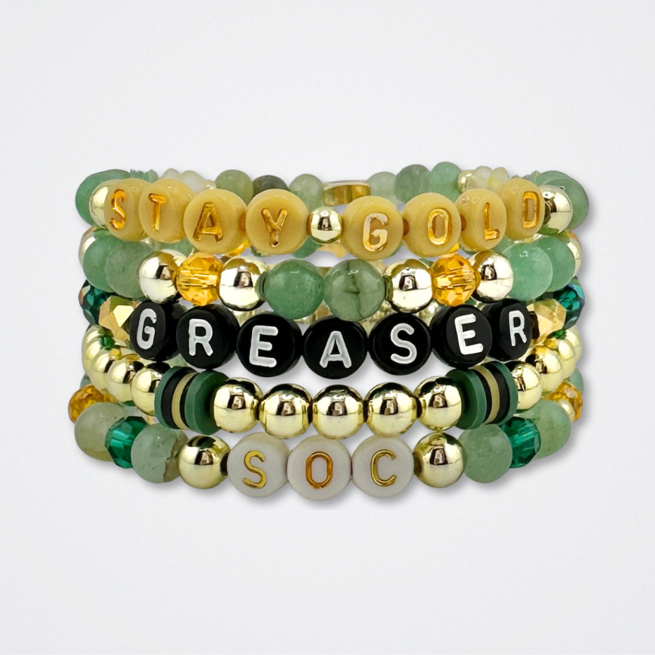 Broadway Stacks STAY GOLD bracelet stack inspired by The Outsiders Musical. Green, yellow and gold beads with letter beads that spell quotes. Displayed on a gray background.