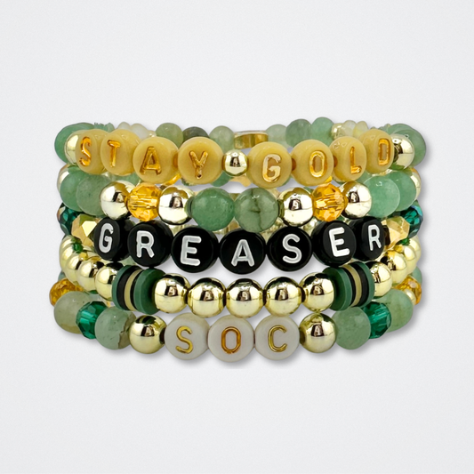 Broadway Stacks STAY GOLD bracelet stack inspired by The Outsiders Musical. Green, yellow and gold beads with letter beads that spell quotes. Displayed on a gray background.