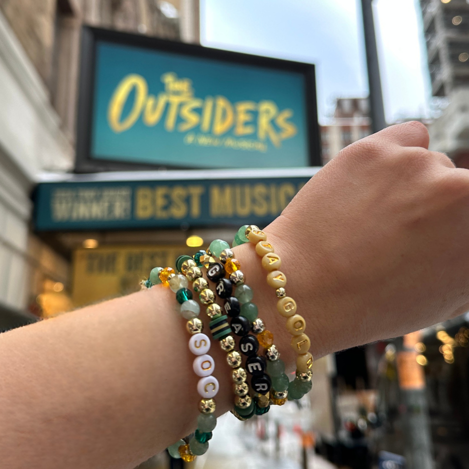 Broadway Stacks STAY GOLD bracelets. Green, yellow and gold beads with letter beads that spell quotes.Bracelets on a wrist in front of The Outsiders Musical marquee.