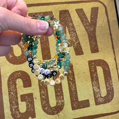 Broadway Stacks STAY GOLD bracelets inspired by The Outsiders Musical. Green, yellow and gold beads with letter beads that spell quotes. Hand holding bracelets in front of a STAY GOLD sign.
