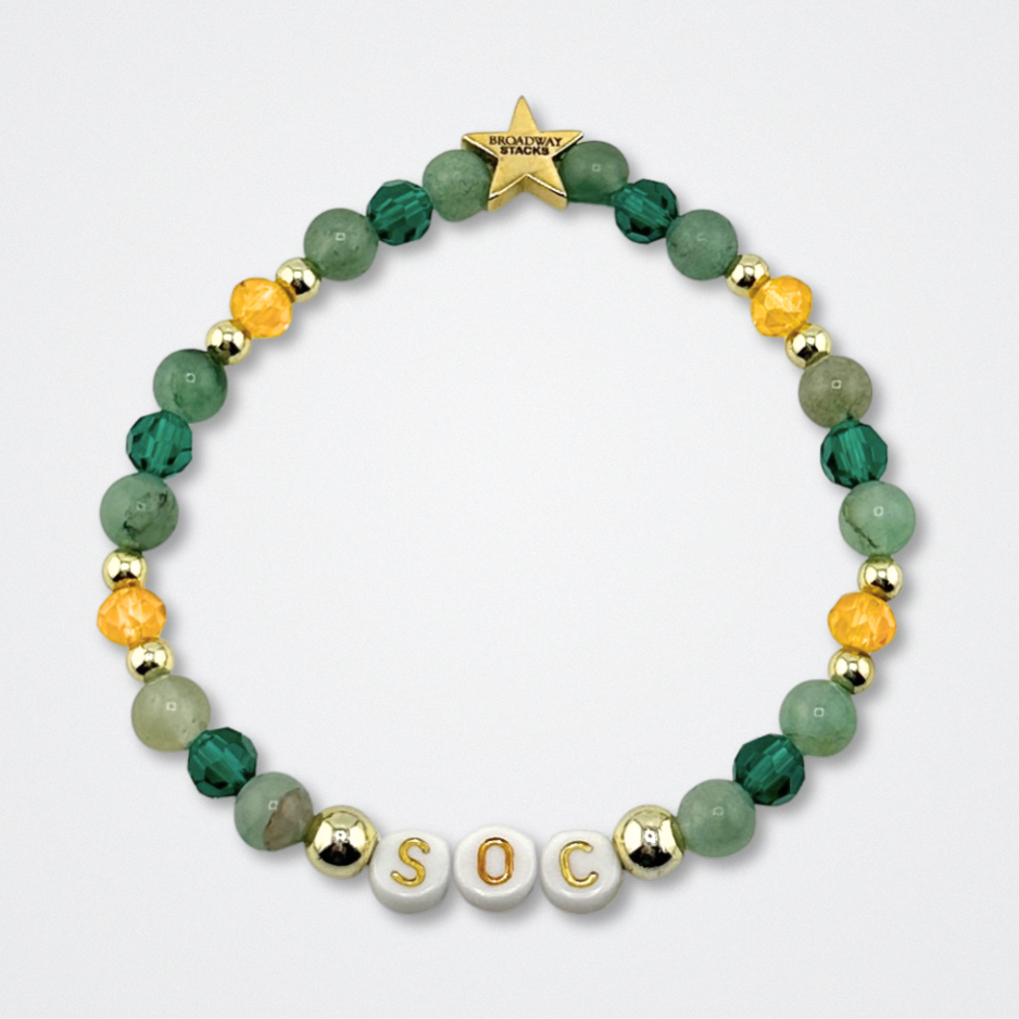 Broadway Stacks STAY GOLD bracelet inspired by The Outsiders Musical. Green and gold beads with letter beads that spell SOC. Logo star bead to remind you that YOU SHINE. Displayed on a gray background.